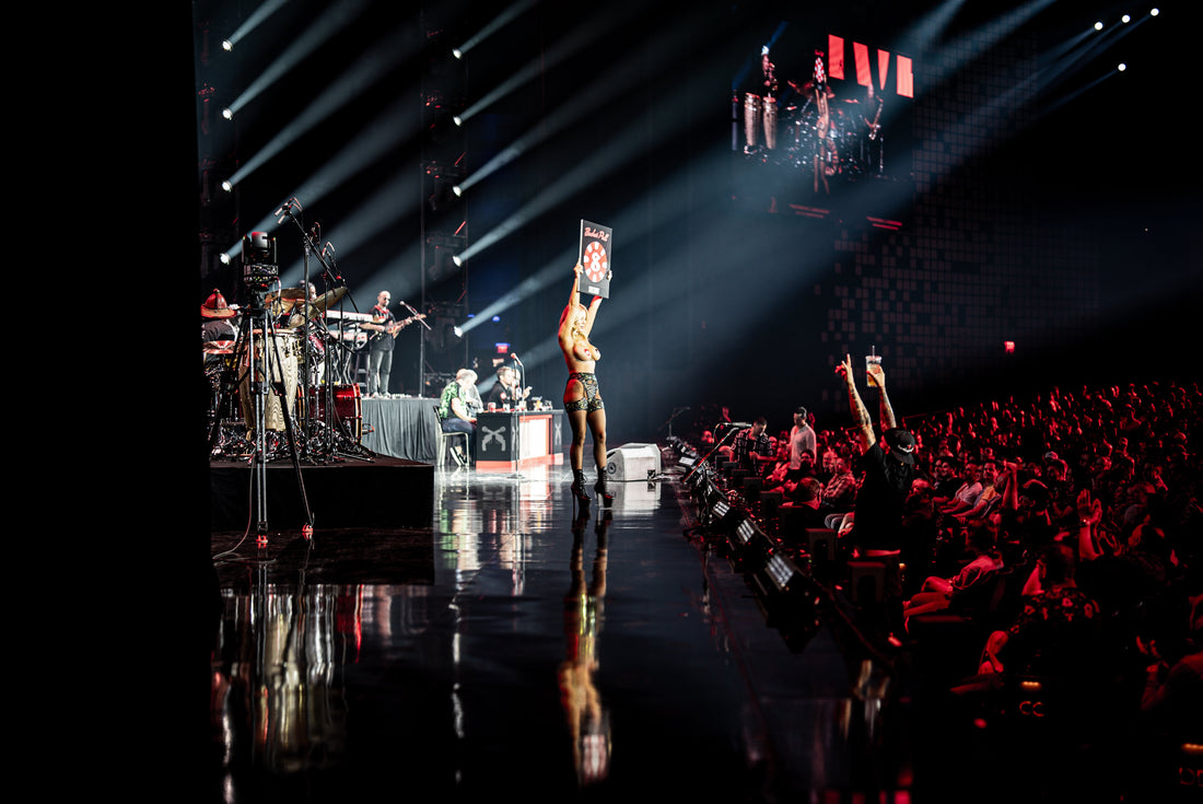Valerie Vaughn holding a sign overhead on the Kill Tony arena stage, with the band and audience under spotlights.